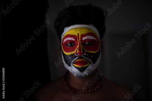 Close up portrait of an Indian brunette man with his face painted by vibrant colors like a tribe standing in front of a studio background. Indian hi fashion.