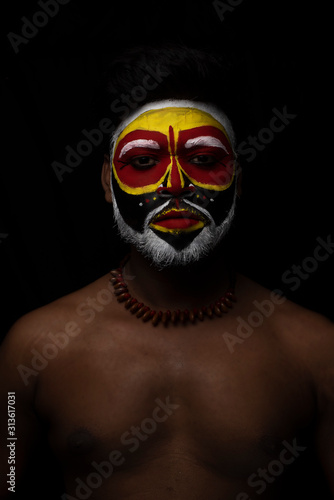 Face of Indian brunette man with his face illuminated and painted by vibrant colors like a tribe standing with candle light in front of a black studio copy space background. Indian hi fashion.