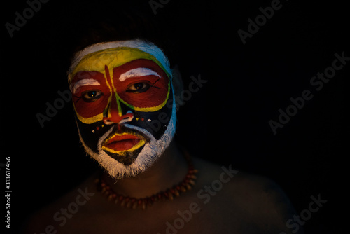 Face of Indian brunette man with his face illuminated and painted by vibrant colors like a tribe standing with candle light in front of a black studio copy space background. Indian hi fashion.