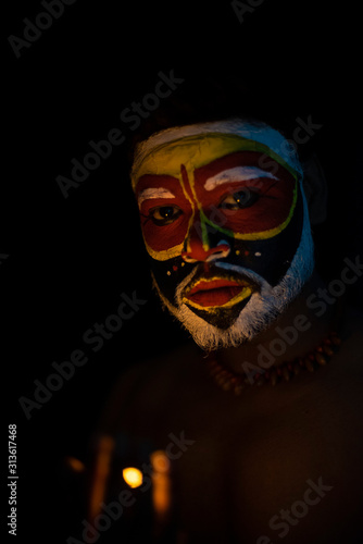 Face of Indian brunette man with his face illuminated and painted by vibrant colors like a tribe standing with candle light in front of a black studio copy space background. Indian hi fashion. © abir