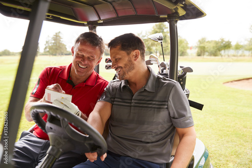 Two Mature Men Playing Golf Marking Scorecard In Buggy Driving Along Course