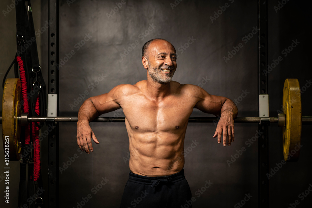Muscular shirtless mature older bodybuilding athlete with balding gray hair leaning against a barbell in a gym smiling at the camera
