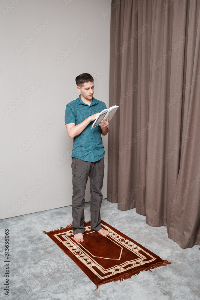 Fototapeta premium A young man prays and recites the muslim Quran on a small portable carpet in the interior. Religion and ritual concept