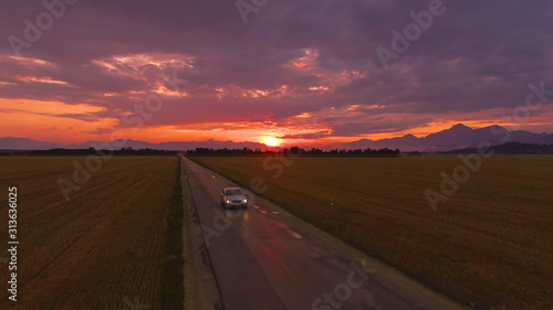 AERIAL: Silver car drives along a scenic country road at breathtaking sunset.