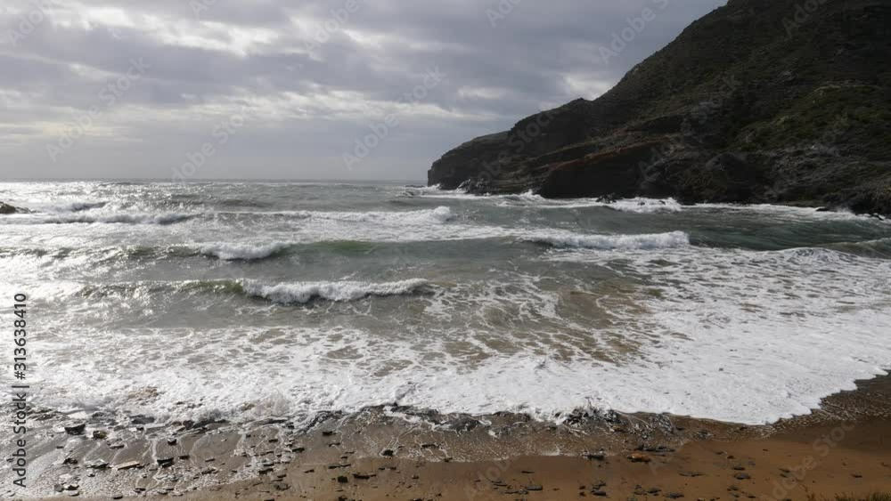 Spanish beach sea waves at cloudy rainy weather. Cala Reona in Cabo de Palos. Mediterranean Sea landscape, coast in Murcia region. Tourist site. Slow motion Spanish beach sea waves at cloudy rainy weather. Cala Reona in Cabo de Palos. Mediterranean Sea landscape, coast in Murcia region. Tourist site. Slow motion