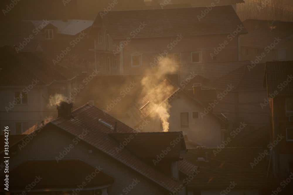 Naklejka Smoking chimneys at roofs of houses emits smoke, smog at sunrise, pollutants enter atmosphere. Environmental disaster. Harmful emissions and exhaust gases into air. Fog, winter day, heating season.
