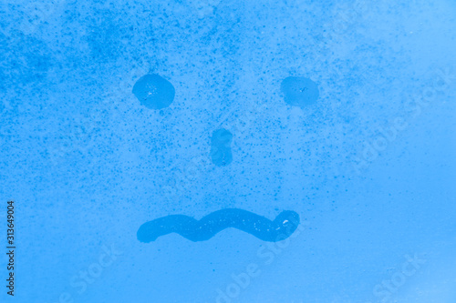 The picture or figure of the scared or tired face on the blue evening or morning window glass with drops 