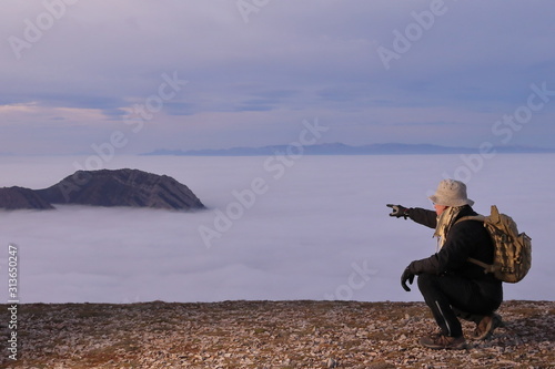  MAN CROUCHED DOWN AT SUMIT SURROUNDED BY CLOUDS POINTING THE HORIZON AT SUNSET