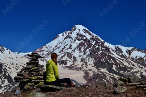 A young girl in yellow sweatshirt sits next to a stone man and looks at the top of the Kazbek mountain covered with glacier. Beautiful sunny day high in the mountains, blue sky. Georgia.