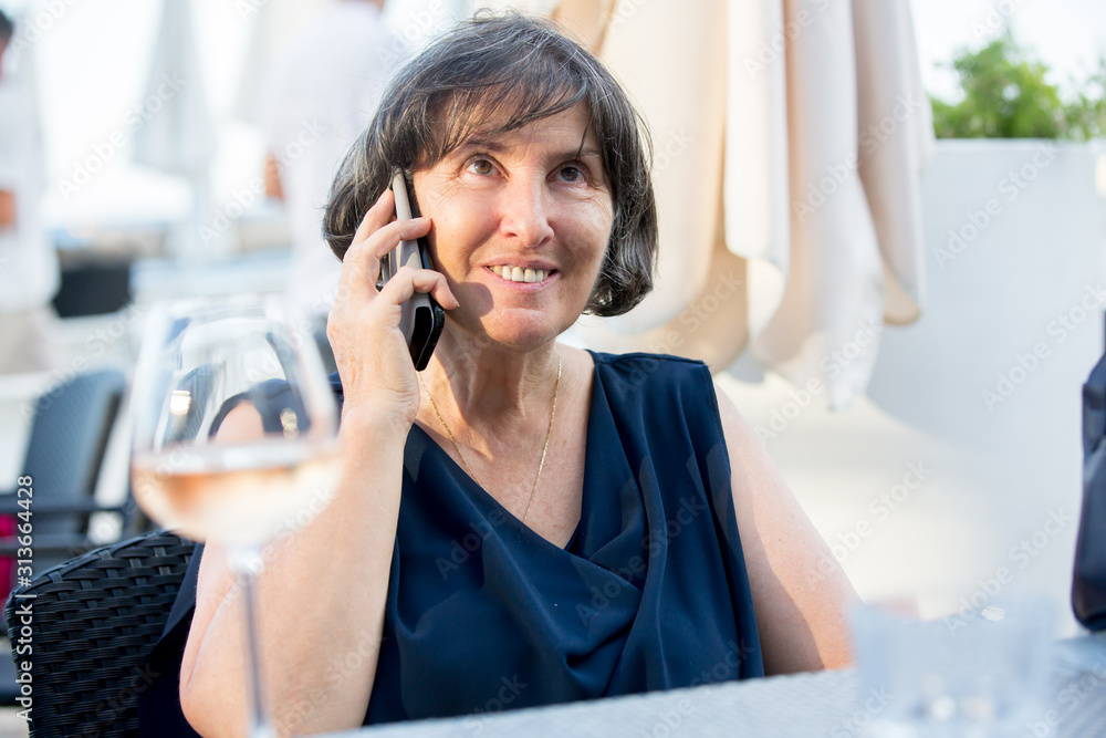 Elderly woman, sitting in beach outdoor restaurant, talking on the phone and drinking rose wine Elderly woman, sitting in beach outdoor restaurant, talking on the phone and drinking rose wine