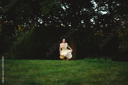 A young woman in a flowing dress runs across a darkened field by woods