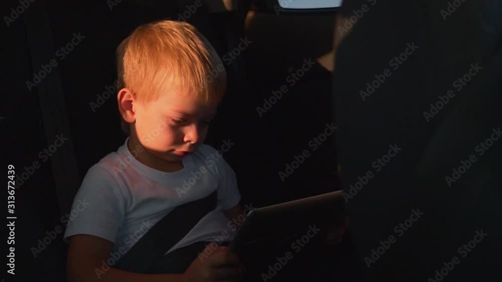 Little boy in the back seat of a car with a tablet in his hands. boy wearing a seat belt. Car travel with a tablet in hand Little boy in the back seat of a car with a tablet in his hands. boy wearing a seat belt. Car travel with a tablet in hand