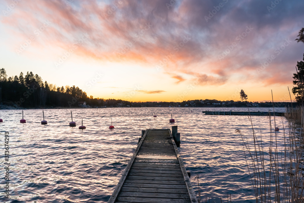Fototapeta premium Old wooden jetty or pier background on the seaside. Sea bay at sunset. Horizon line, skyline with beautiful clouds in the rays of the setting sun. Colorful buoys floating on the surface of the water.