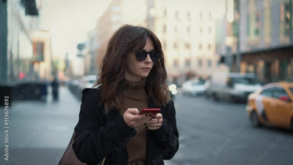 Slow motion of woman in sunglasses with a red phone on background of city road and car passing. Gimbal shot of woman typing in the phone walking along the road Slow motion of woman in sunglasses with a red phone on background of city road and car passing. Gimbal shot of woman typing in the phone walking along the road