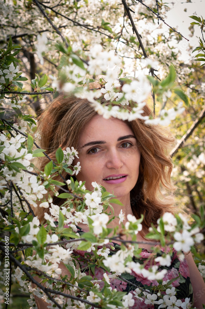 selective focus of beautiful woman with long hair standing behind tree with flowering cherry branches and looking at camera in garden