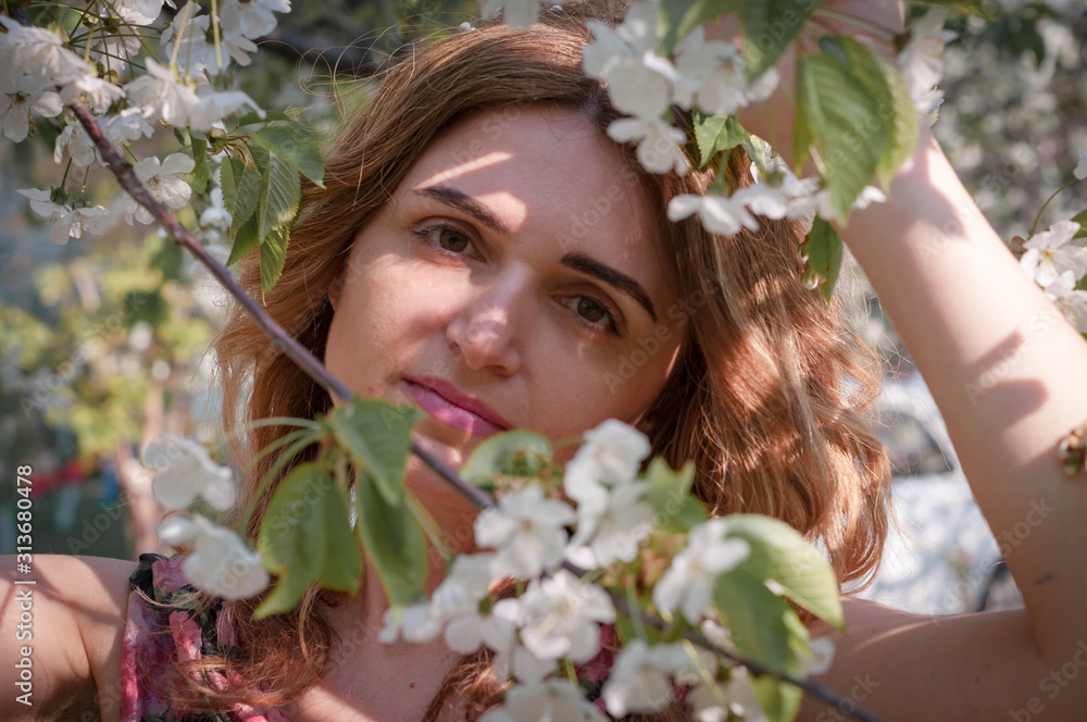 Obraz premium soft focus of portrait of attractive woman with long hair standing behind blooming branches with white flowers and green leaves and looking at camera in garden