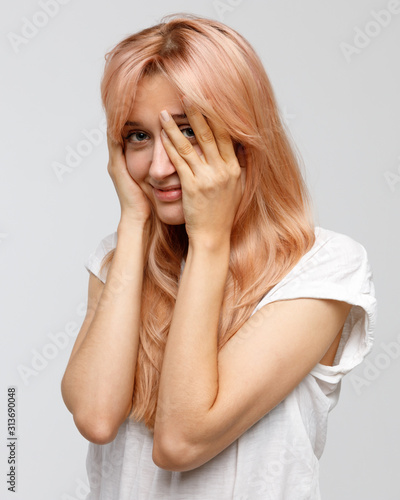 Studio portrait of shy timid woman with long blonde hair covering her face with hand, smiling and peeking through fingers against grey background. Tenderness, sincere emotions. 