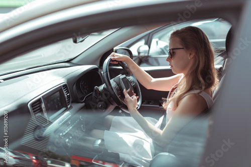 woman in car, sitting behind wheel of a car, reads and writes message in mobile phone, parking mall, traffic jam, in summer in car. Girl in pink dress, automatic transmission.