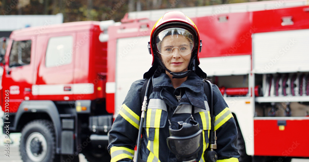 Portrait of young woman firefighter standing near fire truck. Fireman in protective suit with oxygen mask and helmet. Portrait of young woman firefighter standing near fire truck. Fireman in protective suit with oxygen mask and helmet.