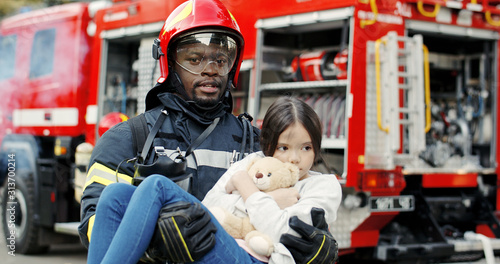 Valokuva Portrait of african american brave fireman holds saved girl in his arms standing near fire truck