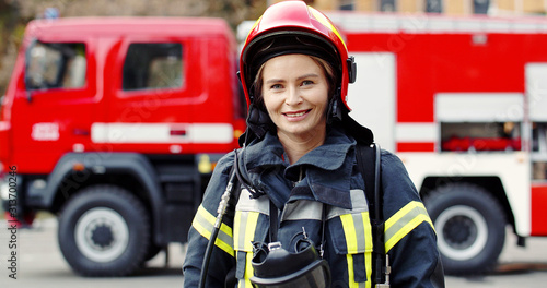 Portrait of young woman firefighter standing near fire truck. Fireman in protective suit with oxygen mask and helmet.