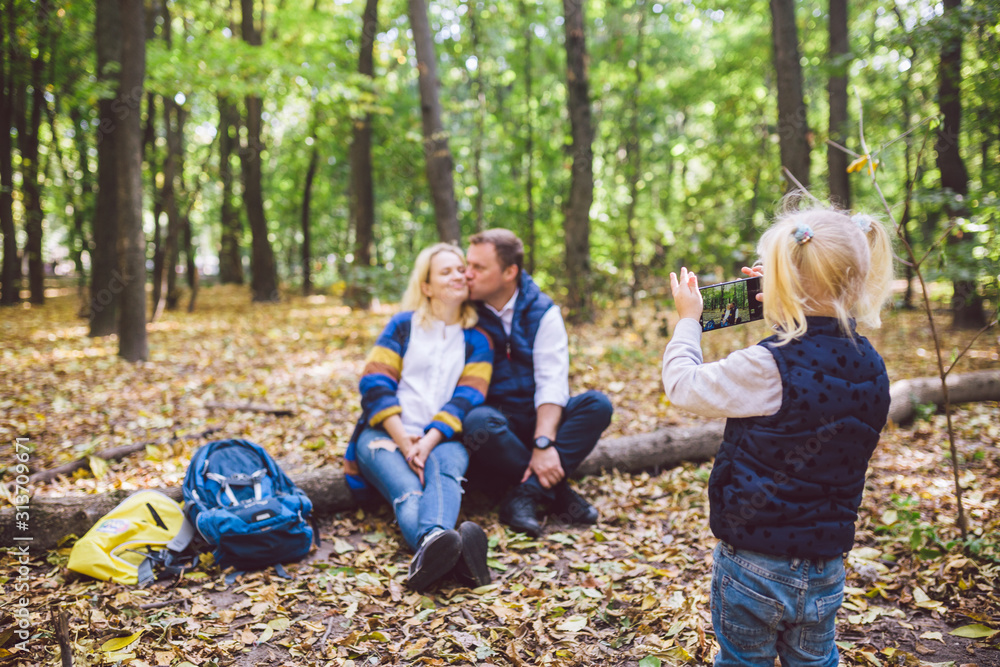 travel, tourism, hike, technology and family concept. Happy child makes photo parents in forest. Mom and dad pose for photo, daughter takes photo on phone. Young family of tourists in wooded area travel, tourism, hike, technology and family concept. Happy child makes photo parents in forest. Mom and dad pose for photo, daughter takes photo on phone. Young family of tourists in wooded area