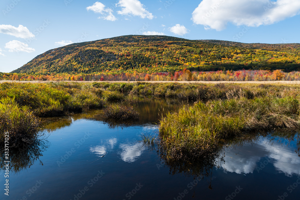 White clouds reflected in blue water below a forest-covered mountain with foliage in fall colors