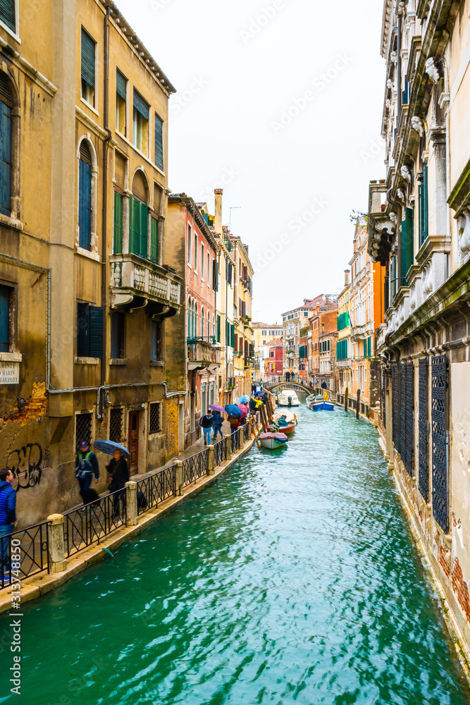 Venice, Italy. Rainy day in the city. Narrow water canal between gothic buildings and a bridge connects islands. People/ Tourists walk with umbrellas. Boats docked and sailing.
