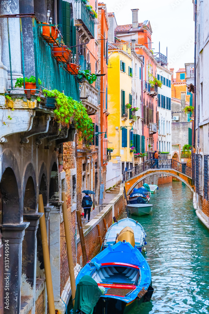 Rainy day in Venice, Italy. Bridge over narrow water canal between colorful gothic Venetian architecture buildings. Boats docked, tourists/ people walking, potted plants hanging from balcony windows.
