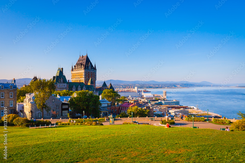 Fototapeta premium Pejzaż widok starego miasta Quebec z budynkami, zielona trawa przed rzeką St Lawrence i błękitne niebo w letni wieczór