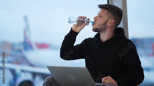 Man drinks water from plastic bottle and works on laptop at airport near window