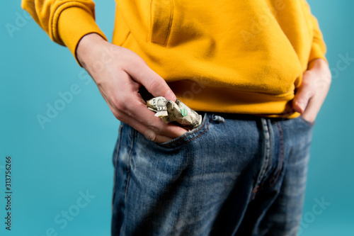 young guy takes out or puts crumpled dollars in his jeans pocket. Teenager in a yellow hoodie on a blue background