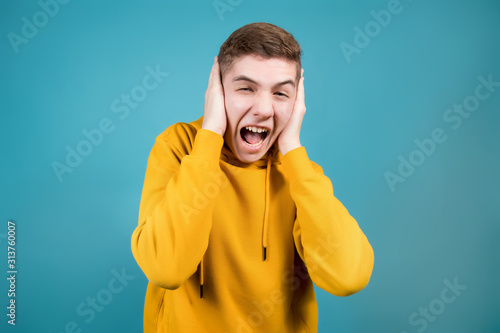 teenager in a yellow sweatshirt on a blue background screams, holding his head in his hands