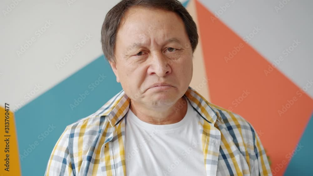 Portrait of unhappy Asian mature man looking at camera with depressed expression standing alone at home feeling lonely and devastated. People and emotions concept. Portrait of unhappy Asian mature man looking at camera with depressed expression standing alone at home feeling lonely and devastated. People and emotions concept.