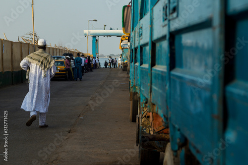A line of cars and trucks waiting in a queue in ferry terminal in Barra. A person is walking towards the sea, with a group of waiting people in the background.