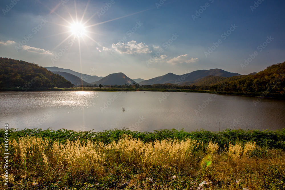 Fototapeta The blurred panoramic nature background of sunlight hitting the lake's surface, grass and wind blowing all the time along the large mountains, ecological beauty and fresh air.