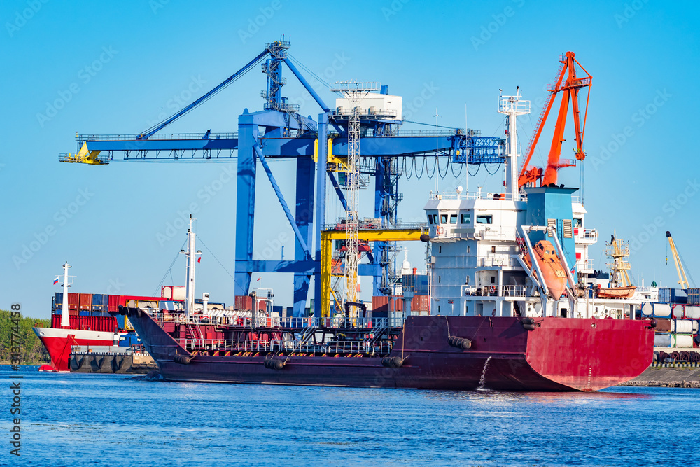 The ship is waiting for loading at the sea commercial port. Ship on the background of cranes. The red ship is moored. Shipping. Freight. Cargo transportation by sea. The ship is waiting for loading at the sea commercial port. Ship on the background of cranes. The red ship is moored. Shipping. Freight. Cargo transportation by sea.