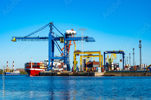 Sea commercial port. Loading container ship. Cranes for cargo transportation. Cargo transportation by sea. The red vessel with containers is preparing to leave the port.