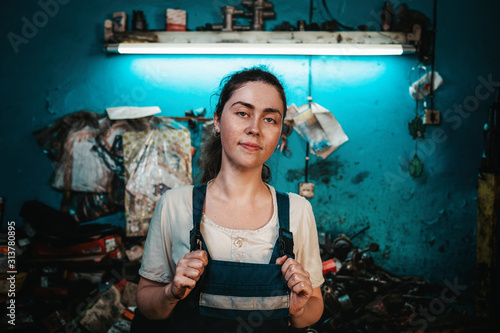 Gender equality. Portrait of a young brunette in uniform, holding hands on the straps of the robe. In the background is an auto repair shop, a blue wall © _KUBE_