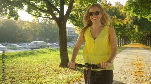 PORTRAIT: Beautiful woman enjoys an electric scooter ride along a sunlit avenue. © helivideo