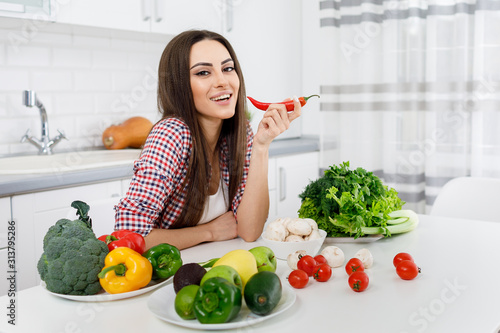 Portrait of beautiful young brunette posing in a kitchen full of greengrocery as if she is going to eat a chili pepper