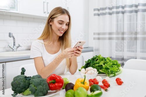 Cute dieting girl using smartphone to check caloricity of greengrocery she has on her kitchen table photo