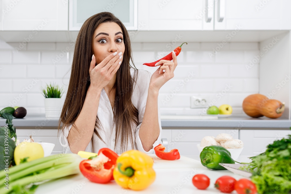 Young brunette model posing with a chili pepper in a light kitchen and covering her mouth with hand Young brunette model posing with a chili pepper in a light kitchen and covering her mouth with hand