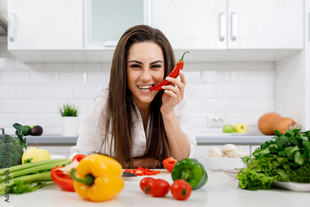Smiling skittish brunette sitting at kitchen table full of vegetables and biting a chili pepper tip