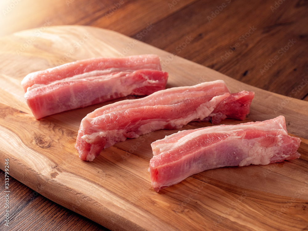 Three raw pork belly slices on a wooden cutting board, Dark wood table surface.