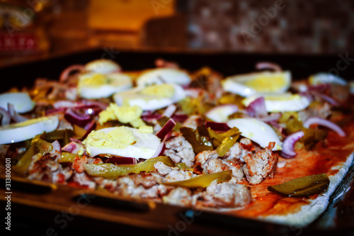 On a black baking sheet is a pizza made from fresh ingredients: dough, ketchup, pickles, minced meat, boiled egg, onions