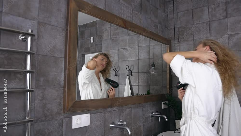 Attractive blonde woman in white bathrobe with long curly hair drying hair in front of mirror in bathroom at home Attractive blonde woman in white bathrobe with long curly hair drying hair in front of mirror in bathroom at home