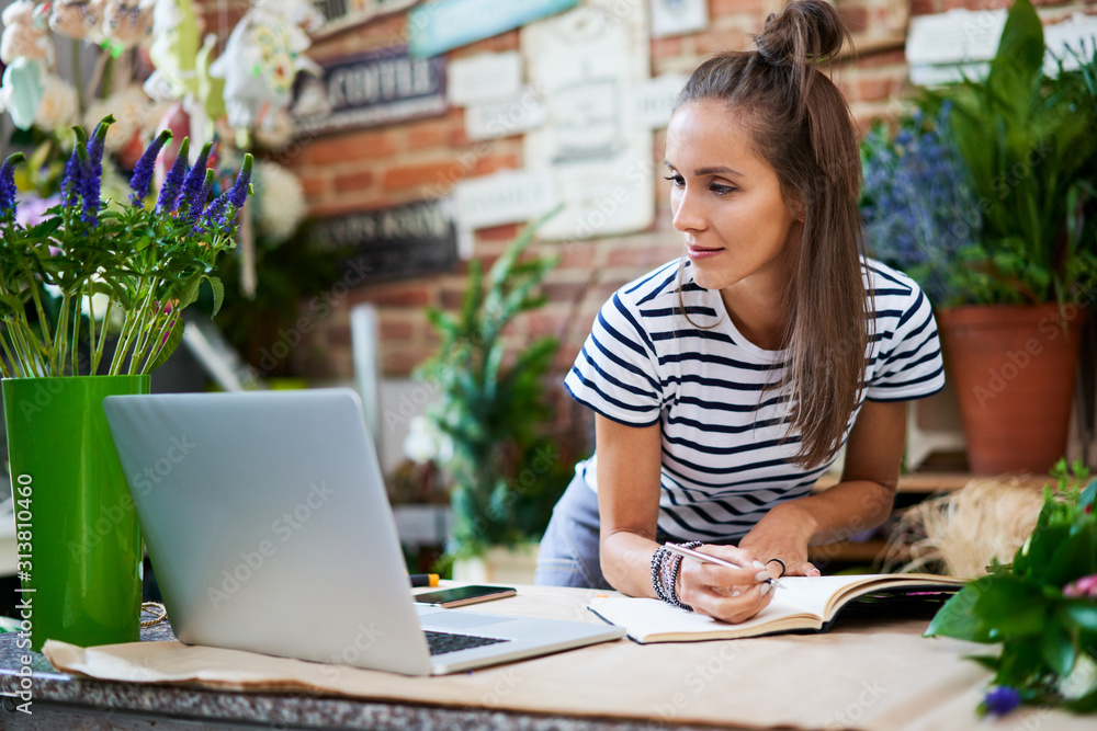 Young florist writing in notebook and looking at laptop while leaning on counter Young florist writing in notebook and looking at laptop while leaning on counter