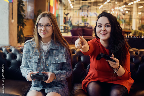 Two girlfriends play video games, holding joysticks in their hands. Console games, addiction, streamers. The brunette points a finger at the screen.