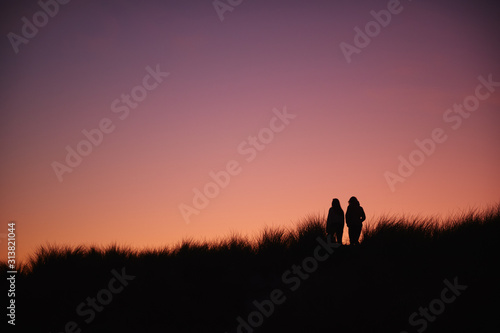 Silhouette Of Two Female Friends Walking Across Dunes Against Setting Sun